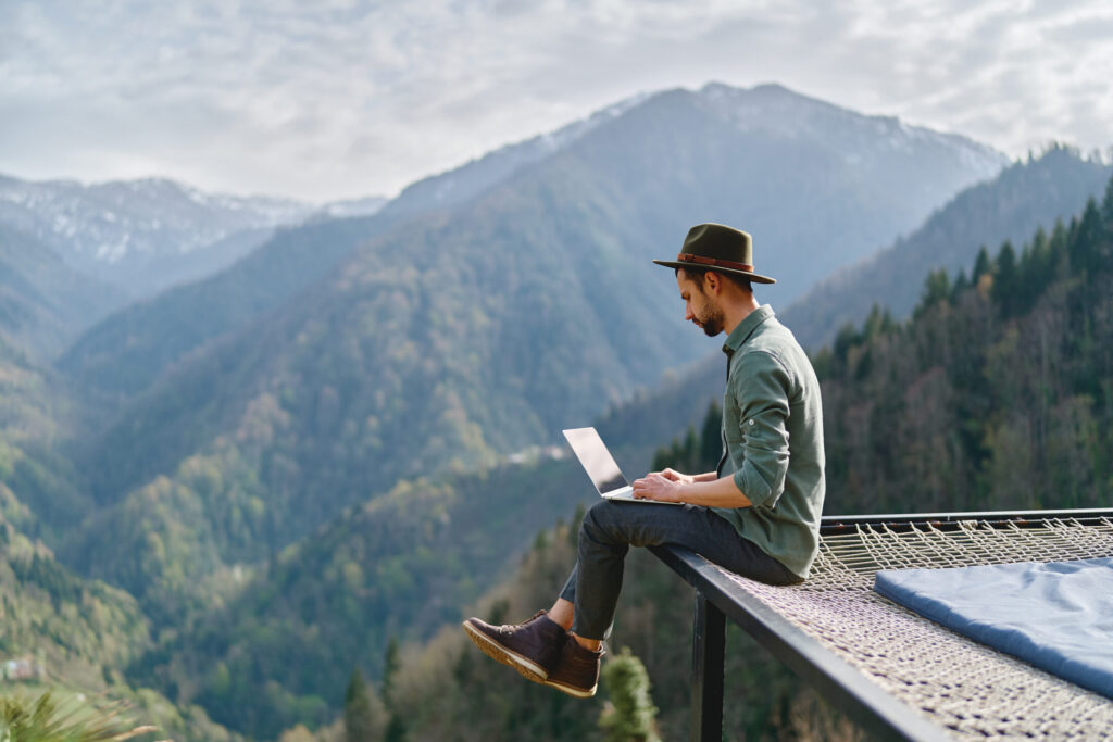Young man with portugal digital nomad visa wearing hat anywhere working online using laptop and enjoying mountains view