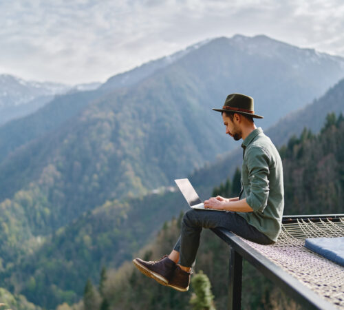 Young man with portugal digital nomad visa wearing hat anywhere working online using laptop and enjoying mountains view